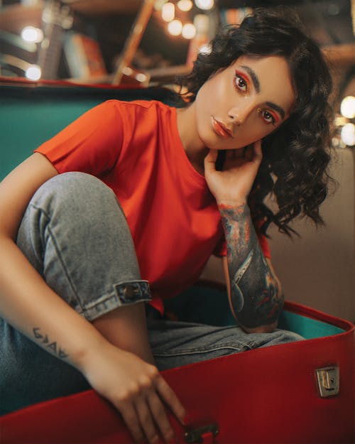 Pensive female in red shirt and jeans with wavy hair and bright makeup looking at camera while chilling in big red old trunk on blurred background