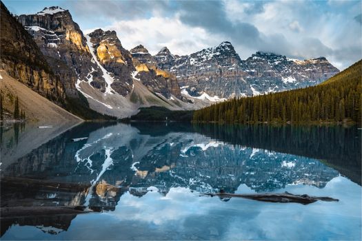 Peaceful lake with clean reflective water located near snowy mountain range and coniferous forest on cloudy day in nature