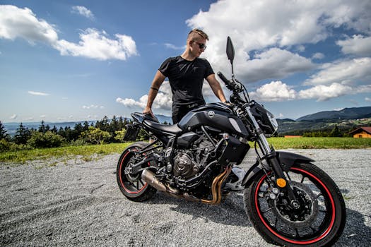 Wide angle of brutal man with haircut standing with black motorcycle on empty rural road in green highlands