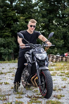 Happy brutal handsome male biker sitting on motorbike and looking at at camera near green trees on sunny day