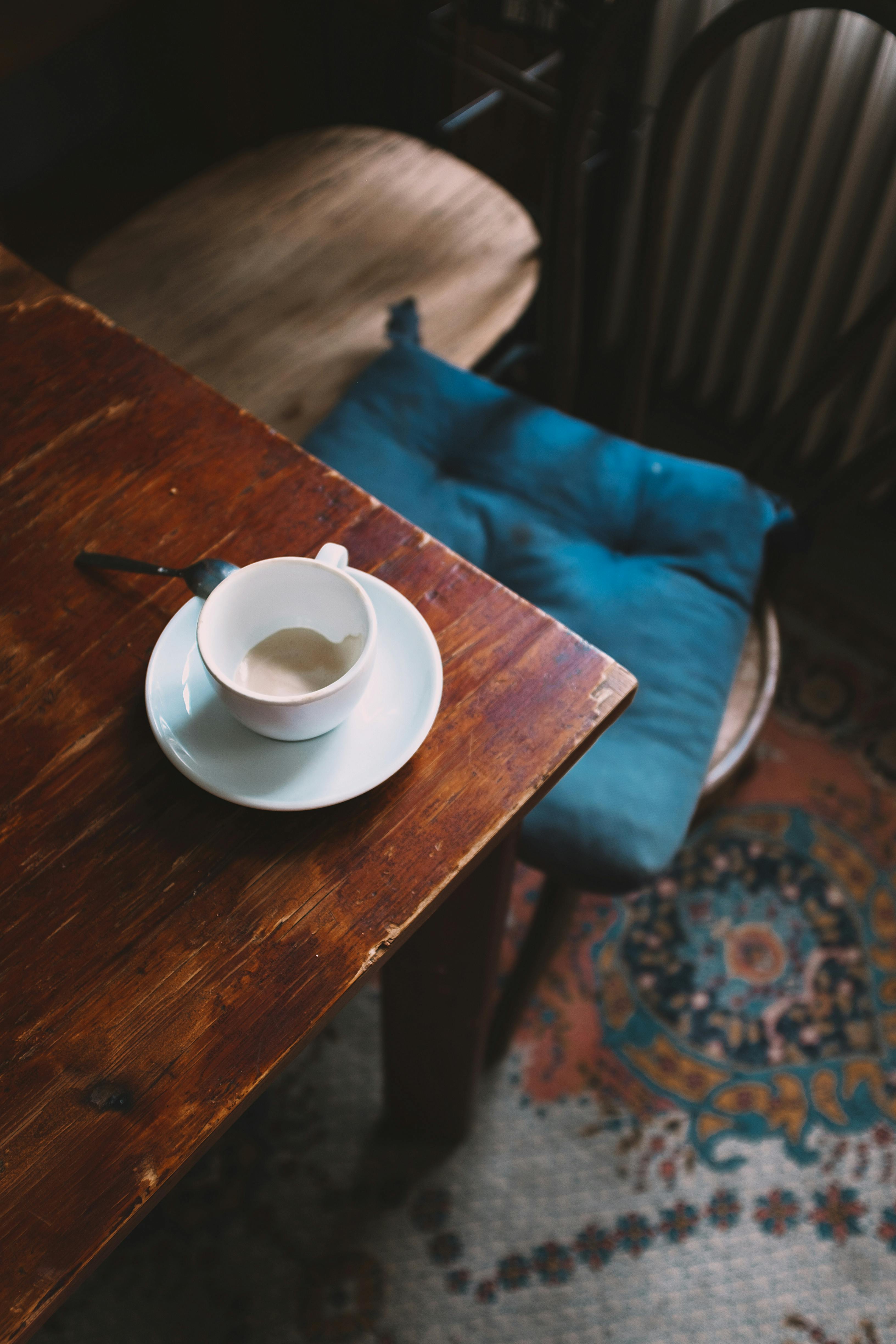 From above of cup of drunk cappuccino placed on weathered wooden table near stool with soft cushion in vintage room