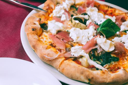 From above of appetizing homemade pizza with prosciutto mozzarella and spinach served on plate and placed on table in cafe