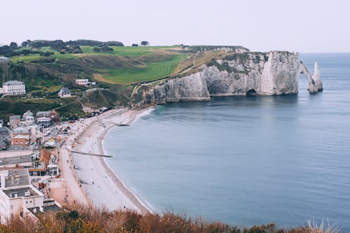 From above of coastal town located on sandy shore near rocky cliff covered with grass and rippling water against cloudless sky