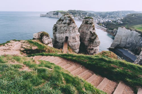 From above of stair surrounded by grass leading to ocean washing rocky cliff on shore near coastal town in nature