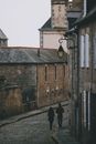 Distant travelers walking on narrow paved walkway near old stone residential houses on street in old town against cloudless sky