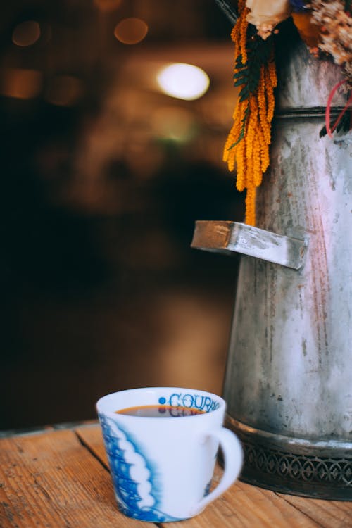 Free Ceramic cup with fresh hot drink placed on wooden table near metal vintage teapot in cozy place on blurred background Stock Photo