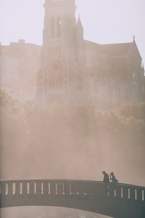 Side view of unrecognizable couple of traveler silhouettes on bridge against old temple and urban building in misty weather