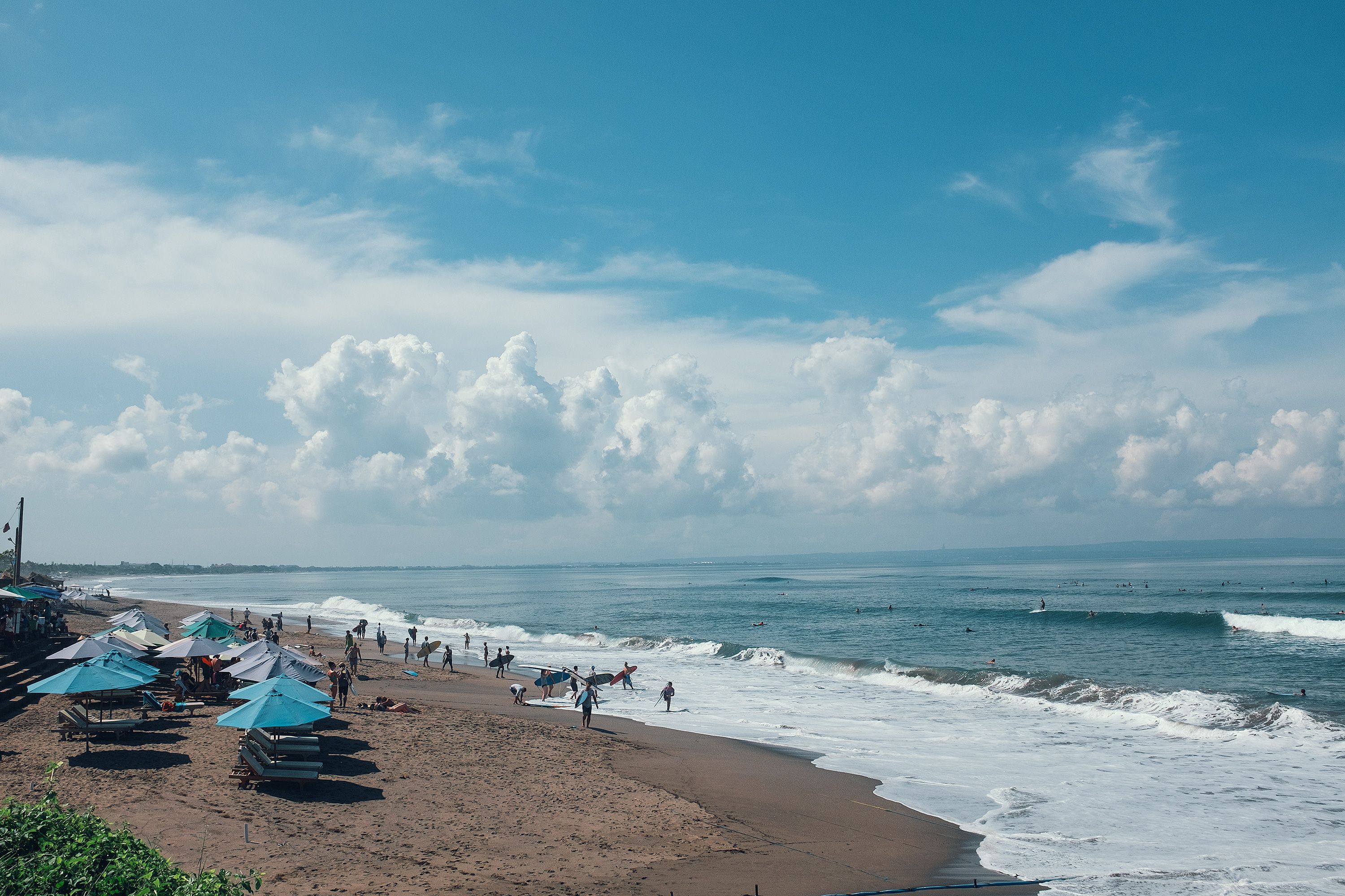 Unrecognizable tourists and athletes with surfboards on sandy beach with parasols near foamy sea under blue cloudy sky
