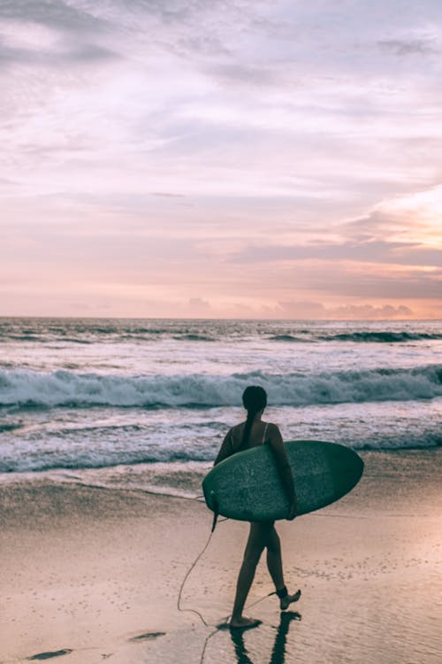 Full body back view of anonymous woman with surfboard walking on wet coast near stormy sea at sunset