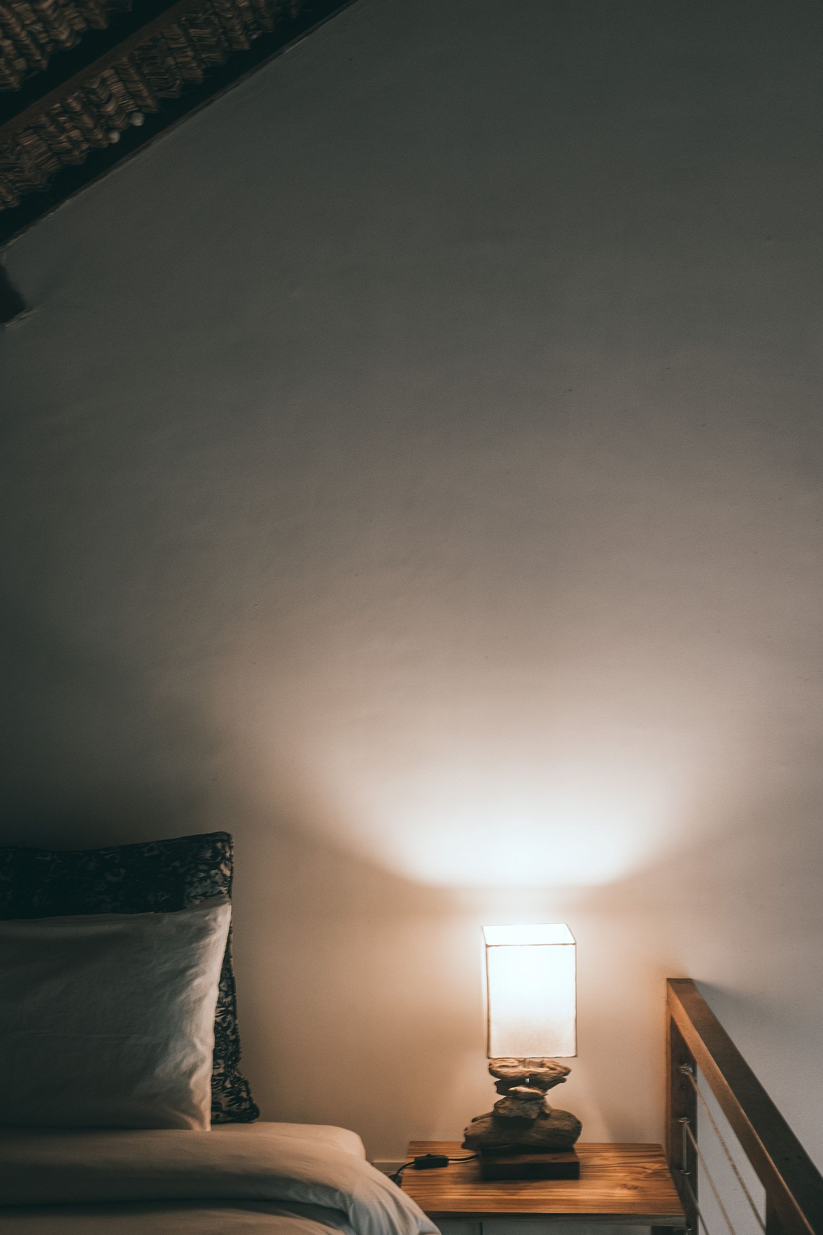 Cama com almofadas ao lado de uma luminária acesa sobre mesa de madeira.