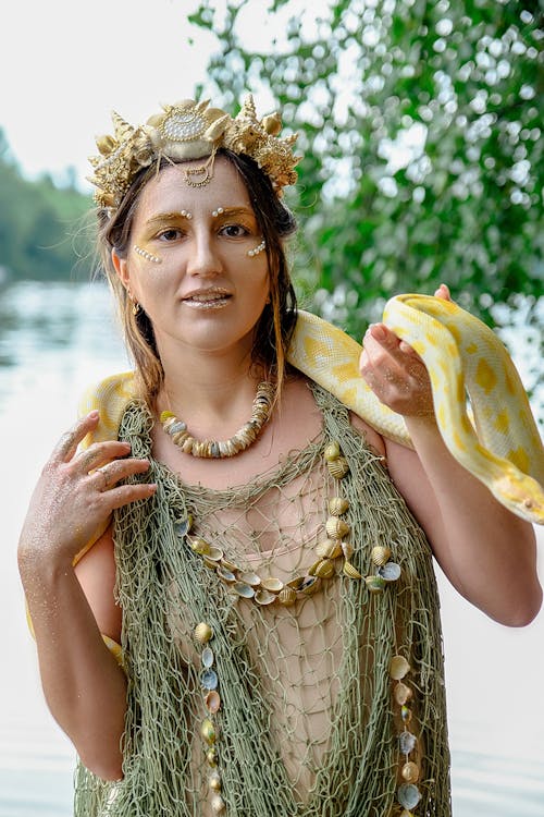 Positive young female model in original costume and makeup with yellow python on neck standing on lake shore and looking at camera