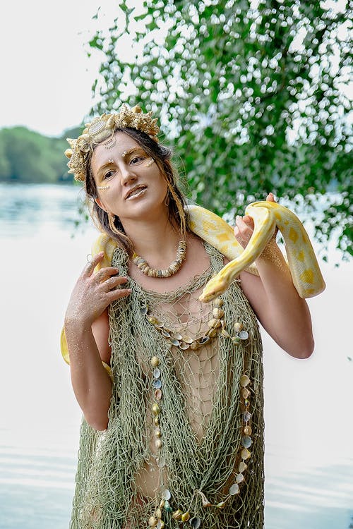 Delighted young pregnant woman in in mesh garment and seashell accessories standing on lake shore with yellow reticulated python on neck and looking away