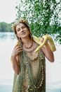 Delighted young pregnant woman in in mesh garment and seashell accessories standing on lake shore with yellow reticulated python on neck and looking away