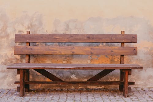 Aged wooden bench placed on paved ground against shabby dirty weathered stone wall of building