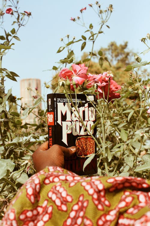 Unrecognizable black person in bright outfit with book in hand near blooming flowers on branches with blurred background in daylight