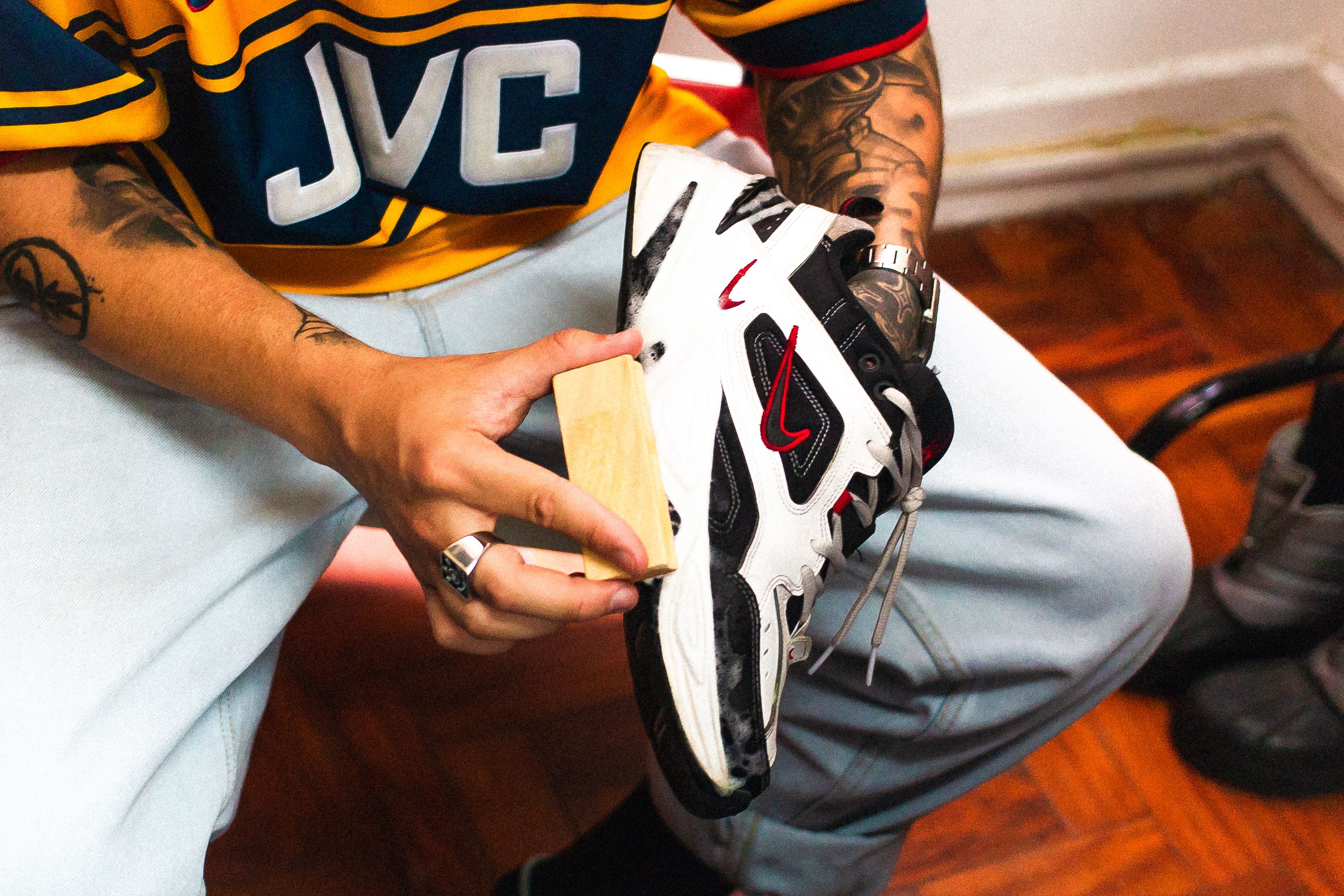 Close-up of a tattooed person cleaning a white sneaker indoors.