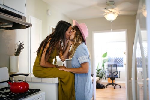 Free A romantic moment captured as two women share a kiss in a bright, cozy kitchen. Stock Photo
