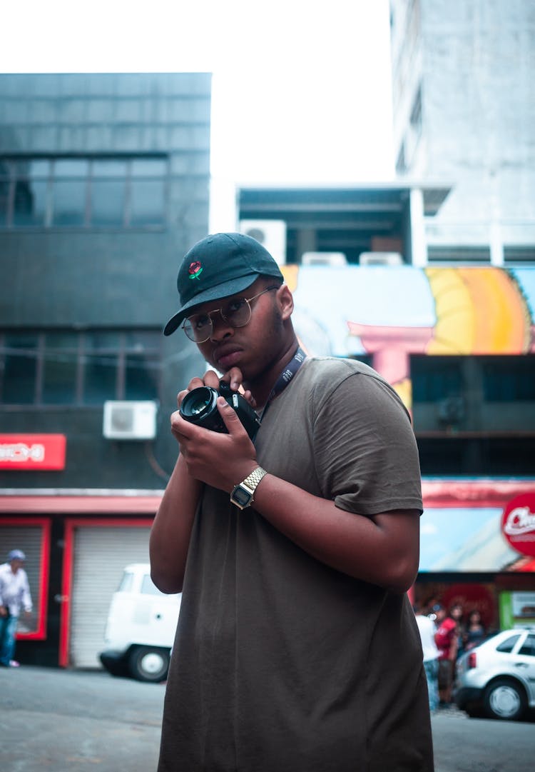 A Young Man With Cap And Eyeglasses Holding A Camera