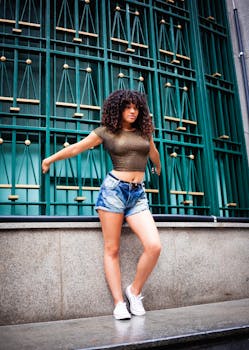 Confident young woman with curly hair in crop top and shorts poses against a patterned urban background.