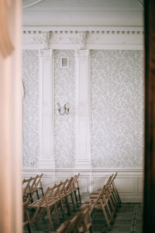 View through doorway of wooden chairs placed in row on tiled floor in room with ornamental wall with white columns