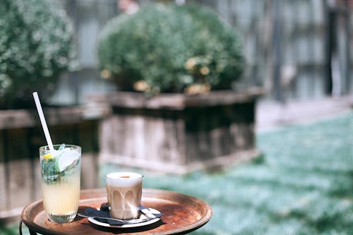 Round tray with glass of delicious cold cocktails and hot coffee placed on street against green bushes on blurred background