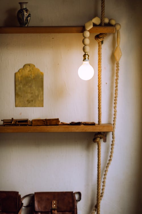 Glowing light bulb illuminating wooden shelves with vase and leather rucksack near wall with stucco in old house