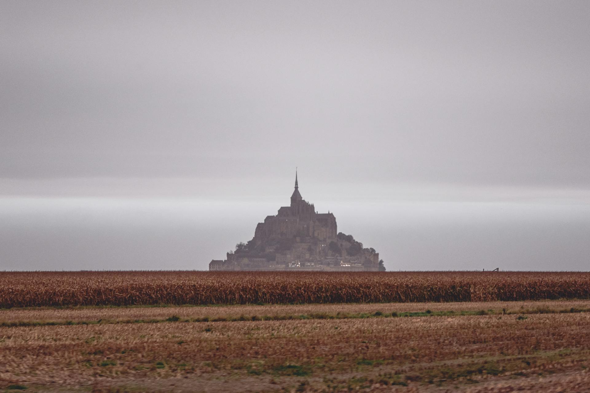 Scenery of distant grand Mon Saint Michel Abbey situated on vast grassy valley under cloudy gloomy sky