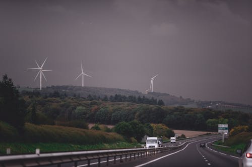 Asphalt highway running through grassy lush valley with modern wind turbine generators under gloomy overcast sky
