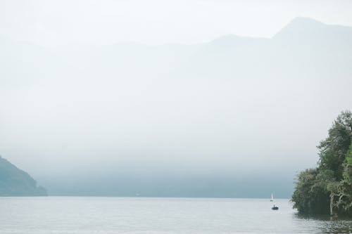 Picturesque scenery of calm lake with boats surrounded with mountains in foggy weather