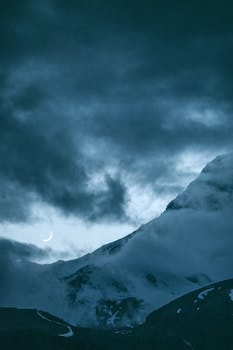 Dramatic view of a snow-capped mountain under a crescent moon and dark clouds.