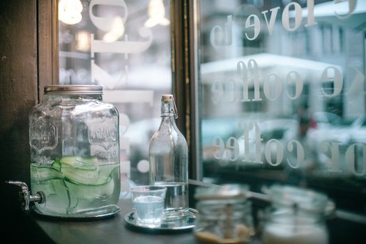 Glass jar of cucumber infuse water and bottle of mineral water with glass placed on wooden table with sugar jars located in urban cafeteria
