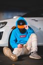 Bearded male driver in stylish outfit browsing smartphone while sitting on road and leaning against car wheel