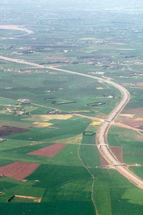 Aerial view of fields covered with green grass near asphalt serpentine road and cottage houses and farms