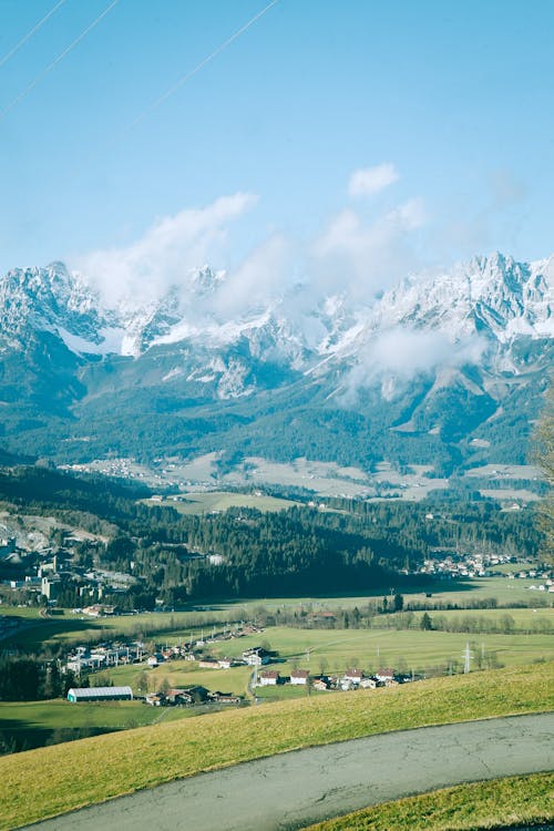 Scenic view of high mount with snow on peaks near grass lawn with rural houses and road under blue cloudy sky