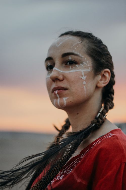 Free A striking portrait of a woman with face paint and braided hair against a sunset backdrop. Stock Photo
