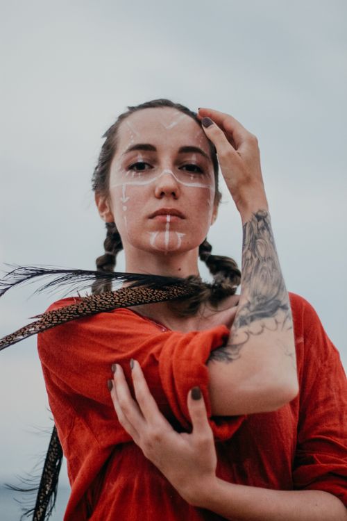 Free A striking portrait of a young woman with face paint, feathers, and a tattoo, wearing a red garment. Stock Photo