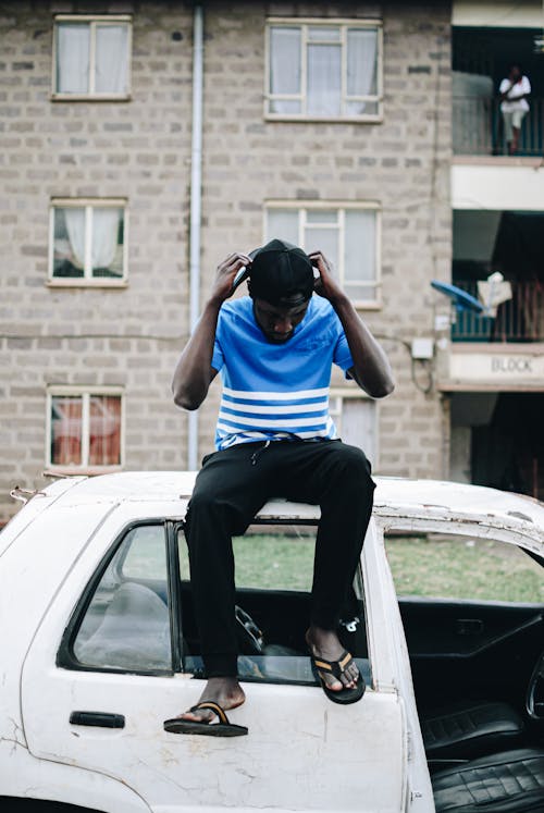 Free Full body of young African American slender male in casual wear touching head and looking down while chilling on roof of old vintage automobile next to multi storey residential building Stock Photo