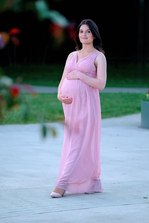 Free Full body of young happy pregnant woman elegant maxi dress embracing belly while walking on alley in park Stock Photo