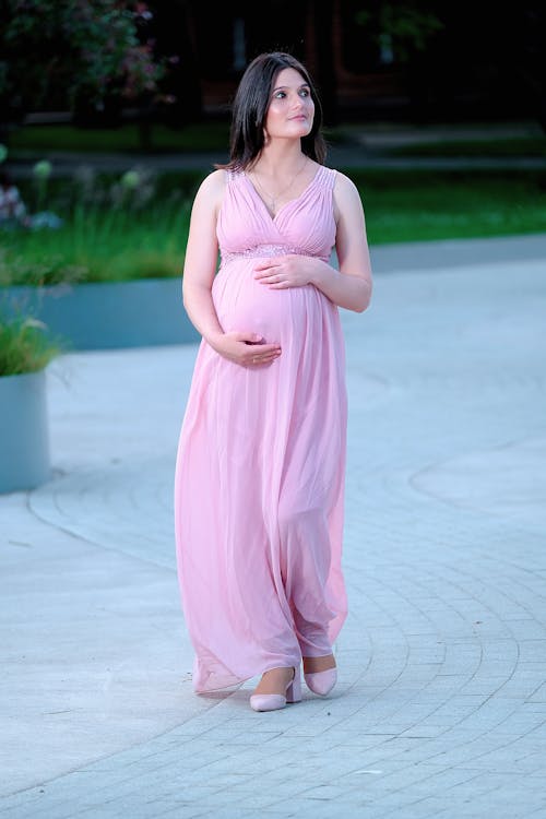 Free Full length of feminine pregnant lady with dark hair in stylish pink dress walking on paved pathway in park and looking away Stock Photo