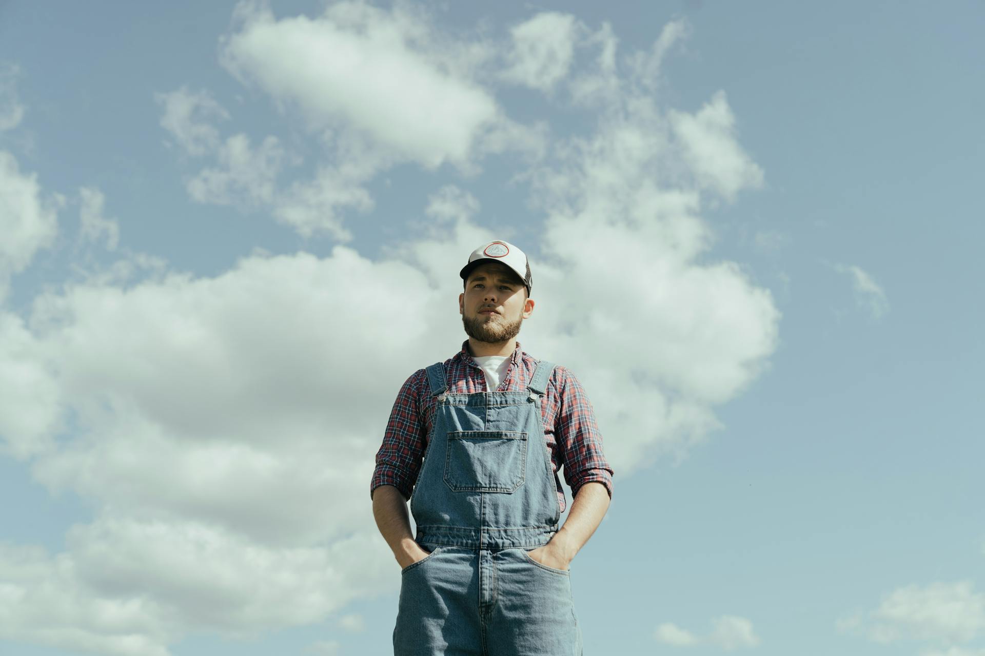 A rural farmer standing confidently in a field, wearing denim overalls and a cap, against a backdrop of a blue sky with clouds.