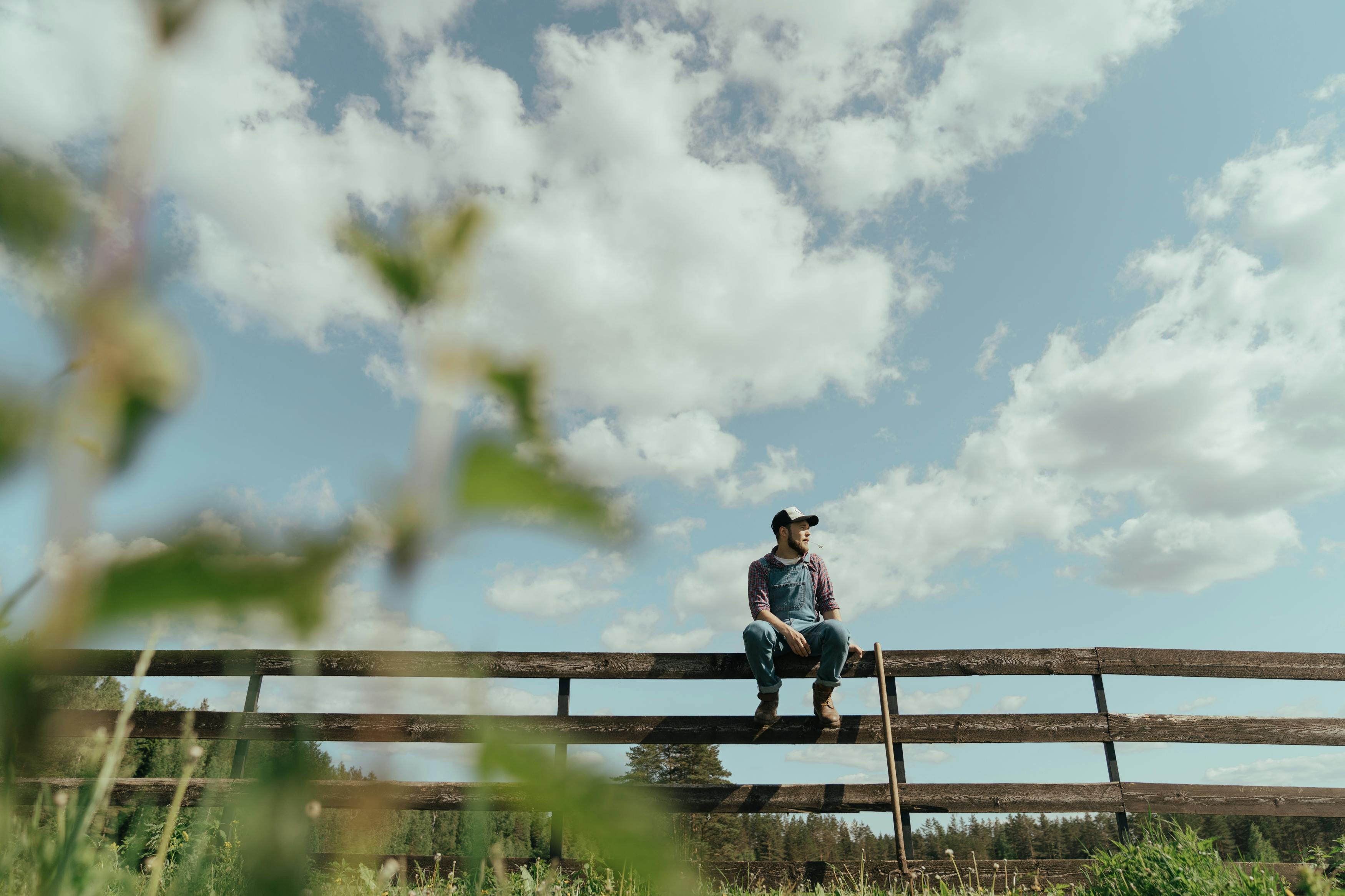 A farmer in overalls sits on a rustic fence under a bright sky, evoking rural charm.