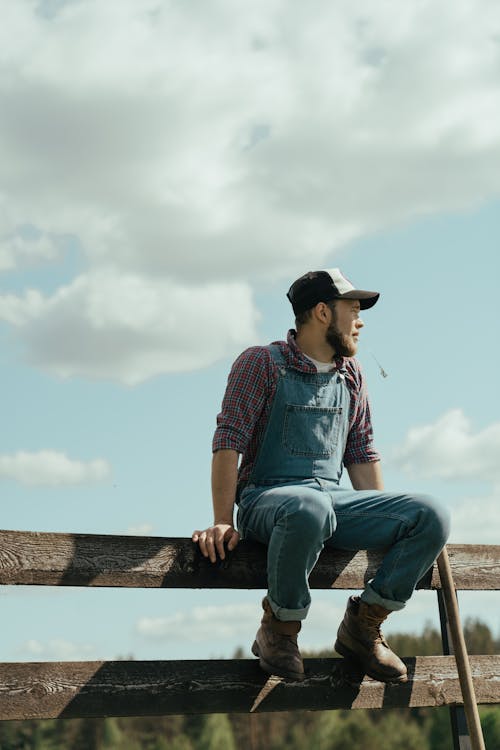 Man in Blue Denim Jeans and Red and Black Plaid Shirt Sitting on Brown Wooden Fence