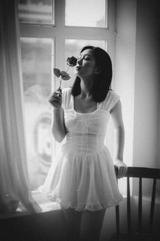 Black and white young Asian woman leaning on chair near window and smelling rose at home