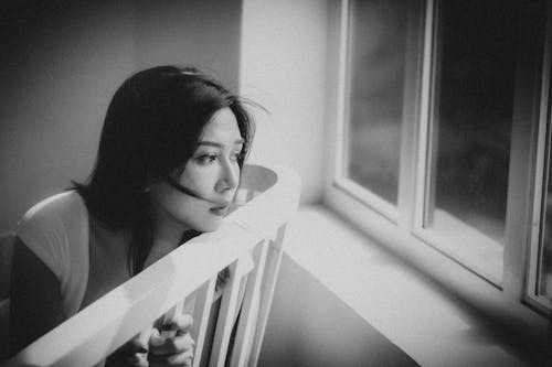 Black and white depressed ethnic female sitting on chair and looking out window at home