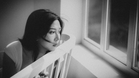 Black and white depressed ethnic female sitting on chair and looking out window at home