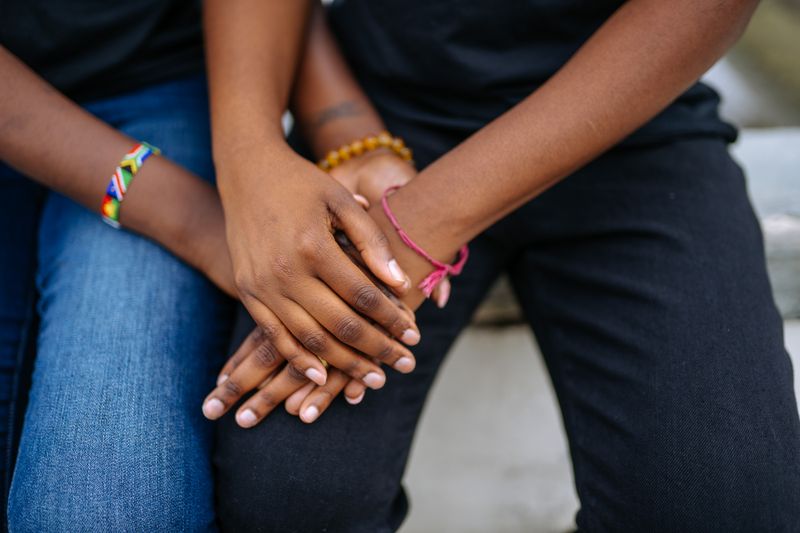 Close-up of two Black adults sitting side by side with their dark brown hands resting clasped on their laps, fingertips and nail beds clearly visible.