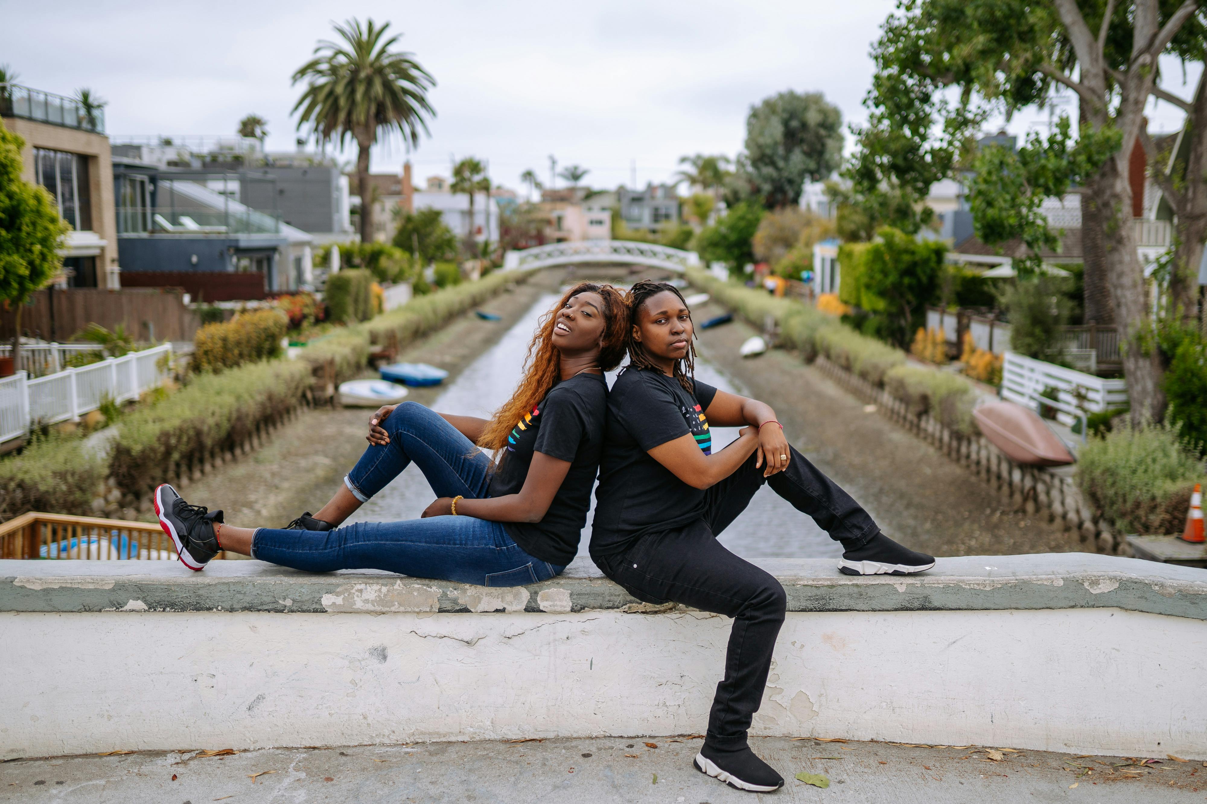 Two women sit back to back wearing matching t-shirts, symbolizing unity and pride by a canal.