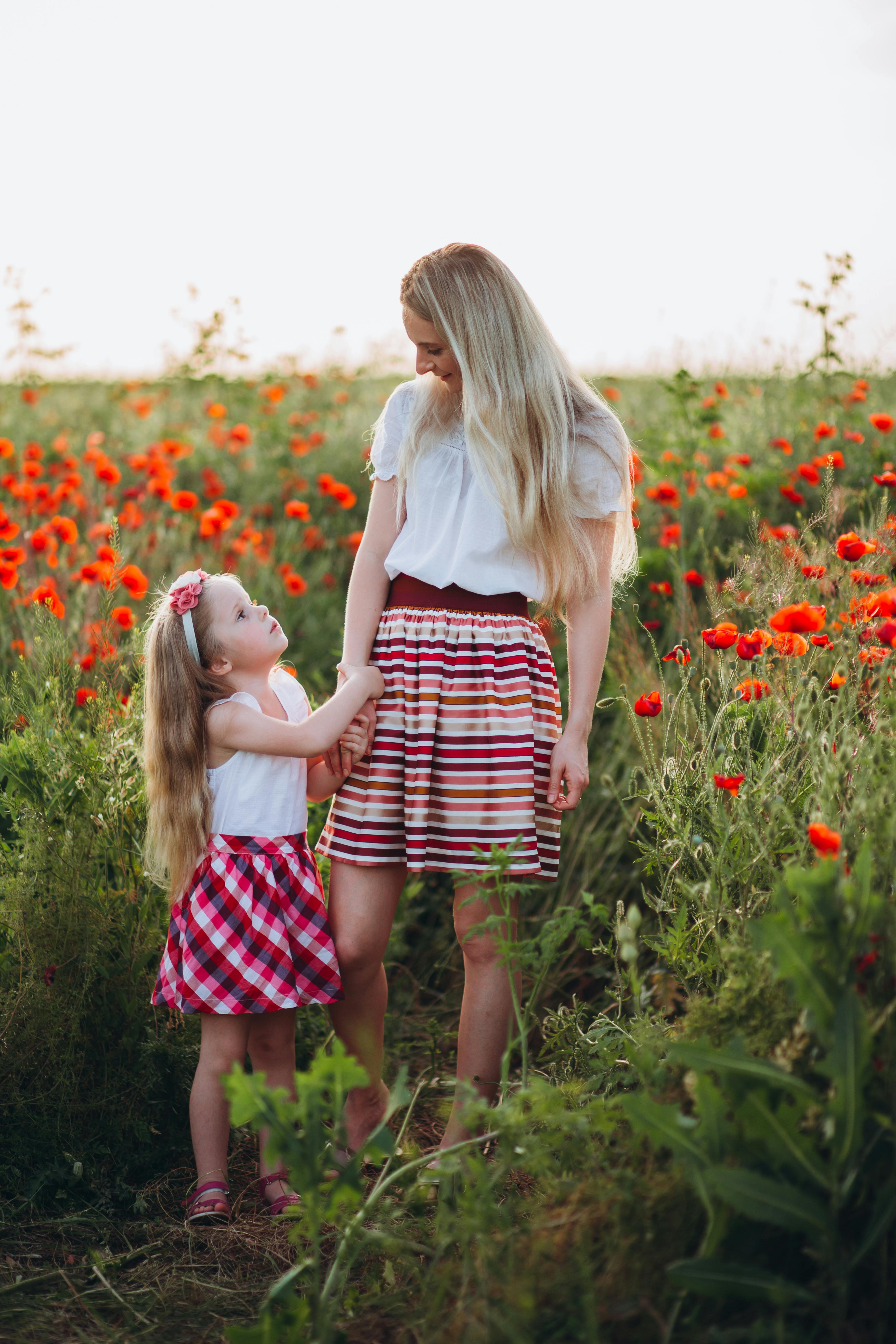Full body of young mother and little daughter holding hands and looking at each other in field of poppies under gray sky