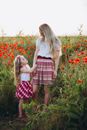 Full body of young mother and little daughter holding hands and looking at each other in field of poppies under gray sky