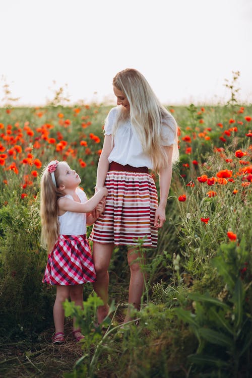 Full body of young mother and little daughter holding hands and looking at each other in field of poppies under gray sky
